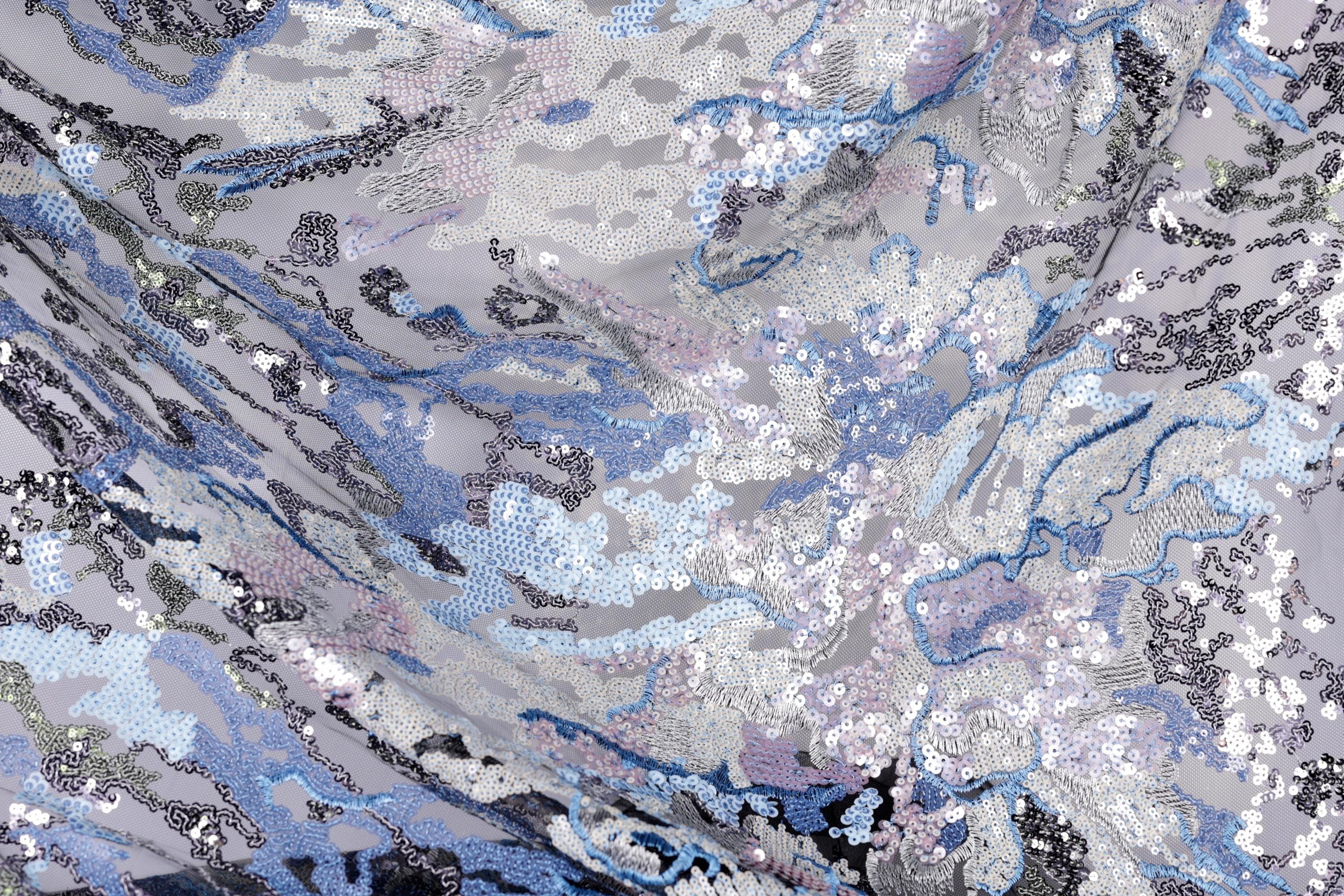 The image is a close-up view of a fabric with a complex, abstract pattern of sequins in various shades of blue, gray, and white. The pattern appears to be a mix of organic and geometric shapes, creating a visually striking and intricate design.