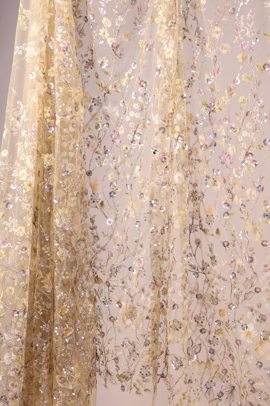 a delicate, floral-patterned fabric with a beige color. The fabric is adorned with numerous small, colorful flowers and leaves, creating a visually appealing and intricate design. The fabric appears to be a sheer or sheer-like material, which adds to its delicate and ethereal appearance.