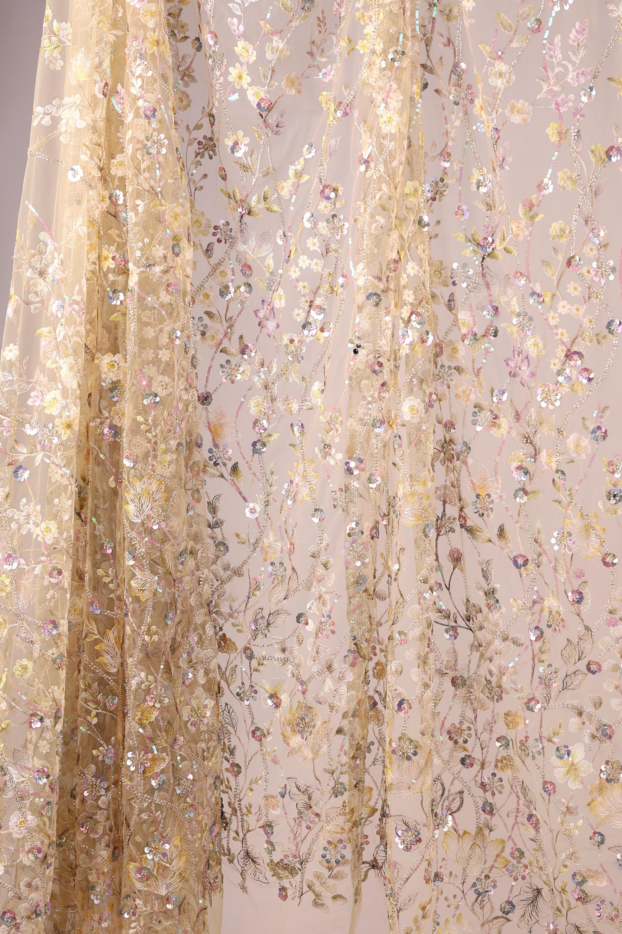 a delicate, floral-patterned fabric with a beige color. The fabric is adorned with numerous small, colorful flowers and leaves, creating a visually appealing and intricate design. The fabric appears to be a sheer or sheer-like material, which adds to its delicate and ethereal appearance.