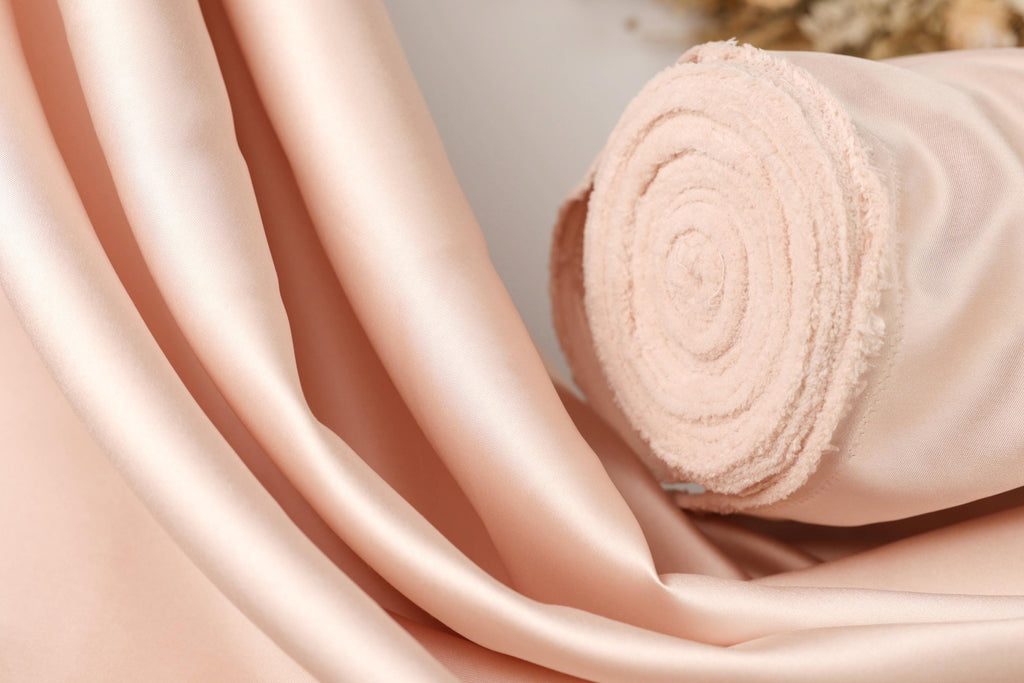 A close-up image of a soft, light pink satin fabric, possibly a satin curtain or fabric, with a rolled-up end visible.