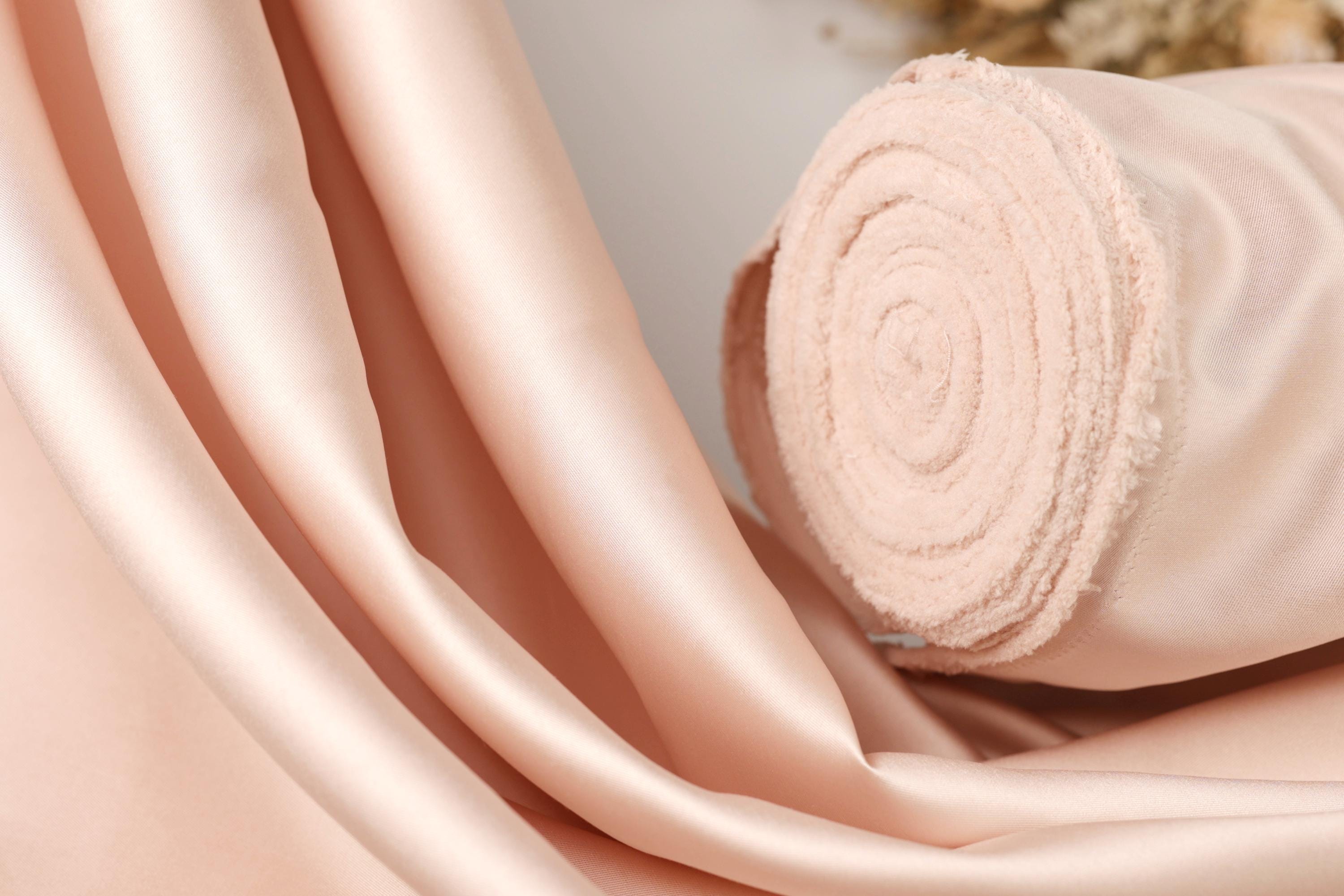 A close-up image of a soft, light pink satin fabric, possibly a satin curtain or fabric, with a rolled-up end visible.