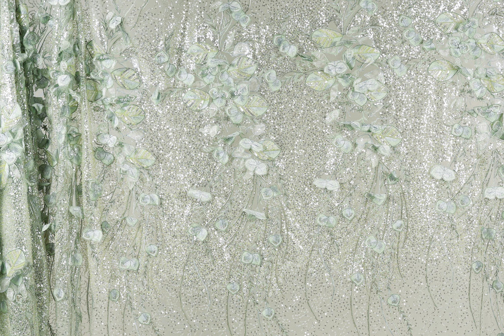 a close-up view of a fabric with a delicate, floral pattern. The fabric appears to be made of a shiny, metallic material, giving it a sparkling, glittery appearance. The pattern consists of numerous small, white flowers or leaves, which are intricately embroidered or printed onto the fabric, creating a visually appealing and detailed design.