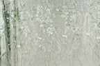 a close-up view of a fabric with a delicate, floral pattern. The fabric appears to be made of a shiny, metallic material, giving it a sparkling, glittery appearance. The pattern consists of numerous small, white flowers or leaves, which are intricately embroidered or printed onto the fabric, creating a visually appealing and detailed design.