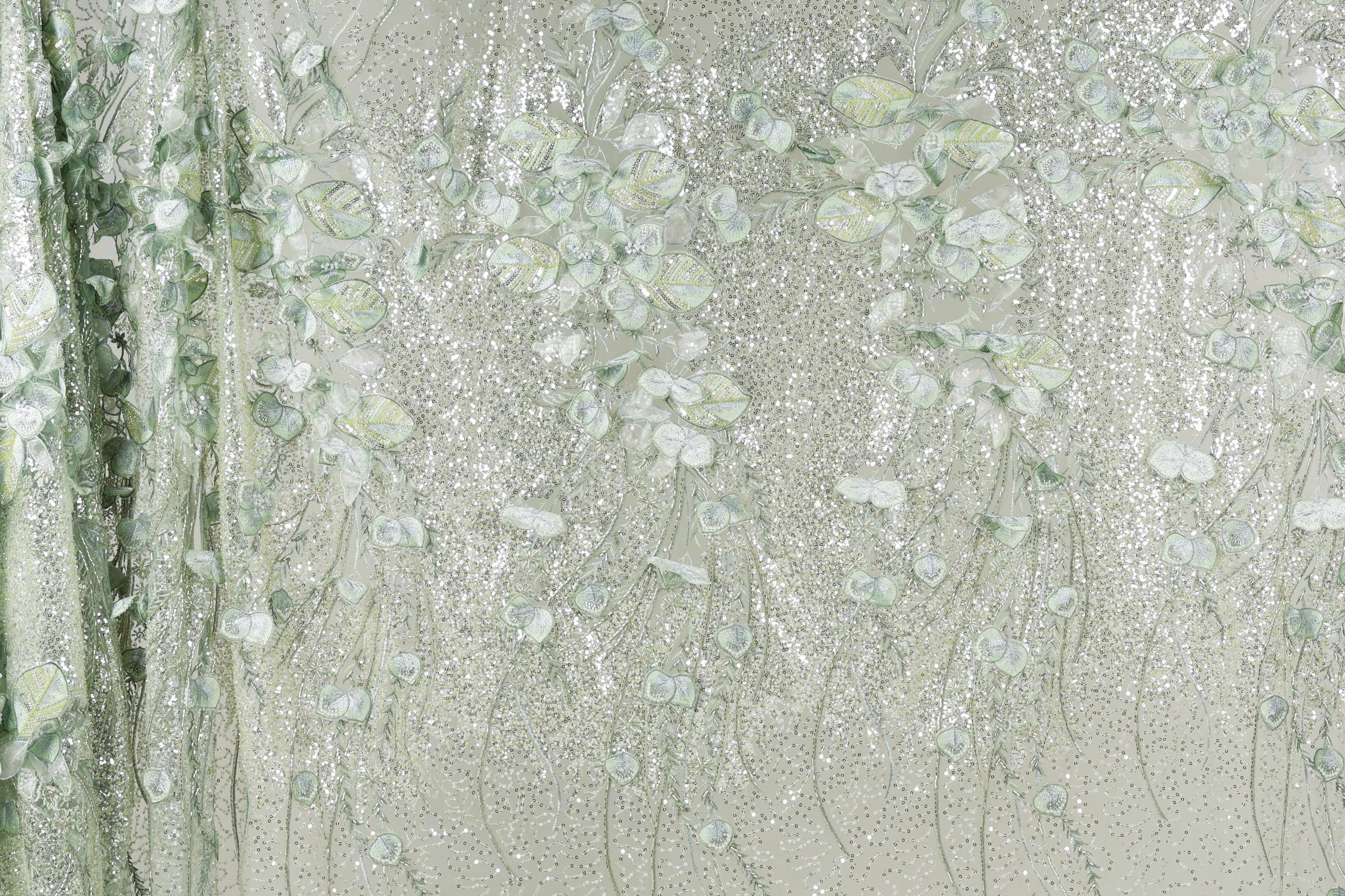 a close-up view of a fabric with a delicate, floral pattern. The fabric appears to be made of a shiny, metallic material, giving it a sparkling, glittery appearance. The pattern consists of numerous small, white flowers or leaves, which are intricately embroidered or printed onto the fabric, creating a visually appealing and detailed design.