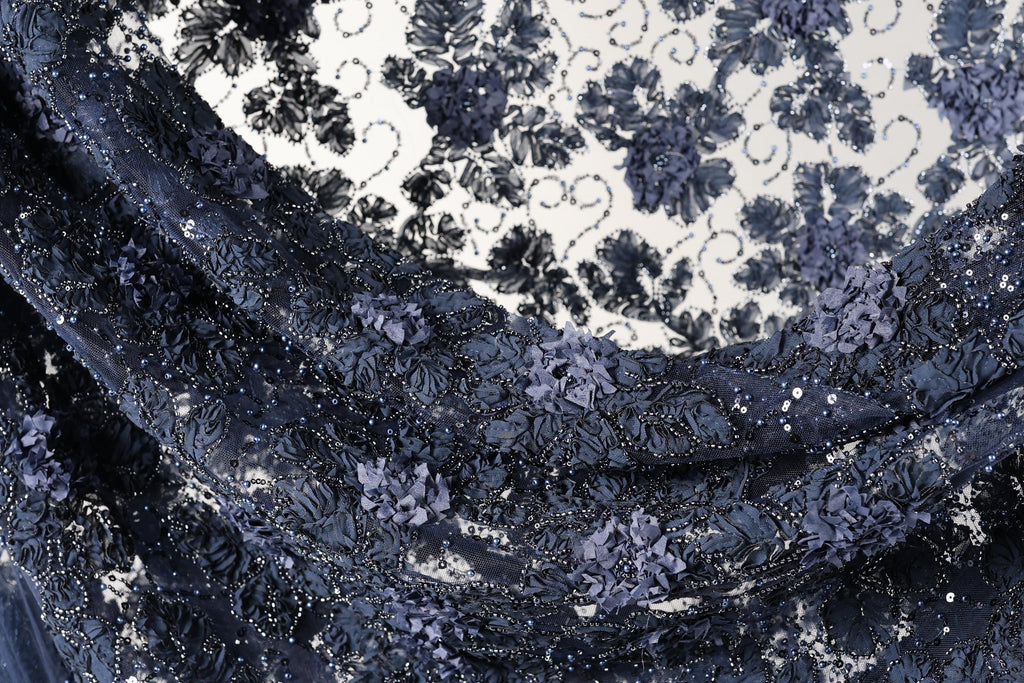 The image is a close-up view of a fabric with a floral pattern, featuring a mix of dark blue and black colors. The fabric appears to be made of lace or a similar material, with intricate details and embellishments.