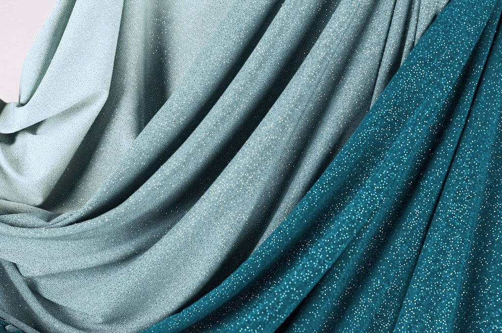 a close-up view of a fabric with a shimmering, sparkly texture in shades of blue and gray. The fabric appears to be made of a shiny, metallic material, giving it a unique and eye-catching appearance.