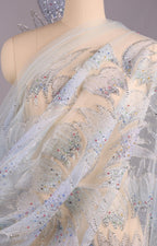 a mannequin wearing a white dress with a sheer, sheer-like fabric and a floral pattern. The dress appears to be made of sheer white fabric with silver embellishments, such as sequins or rhinestones, adding a touch of sparkle and elegance to the design.