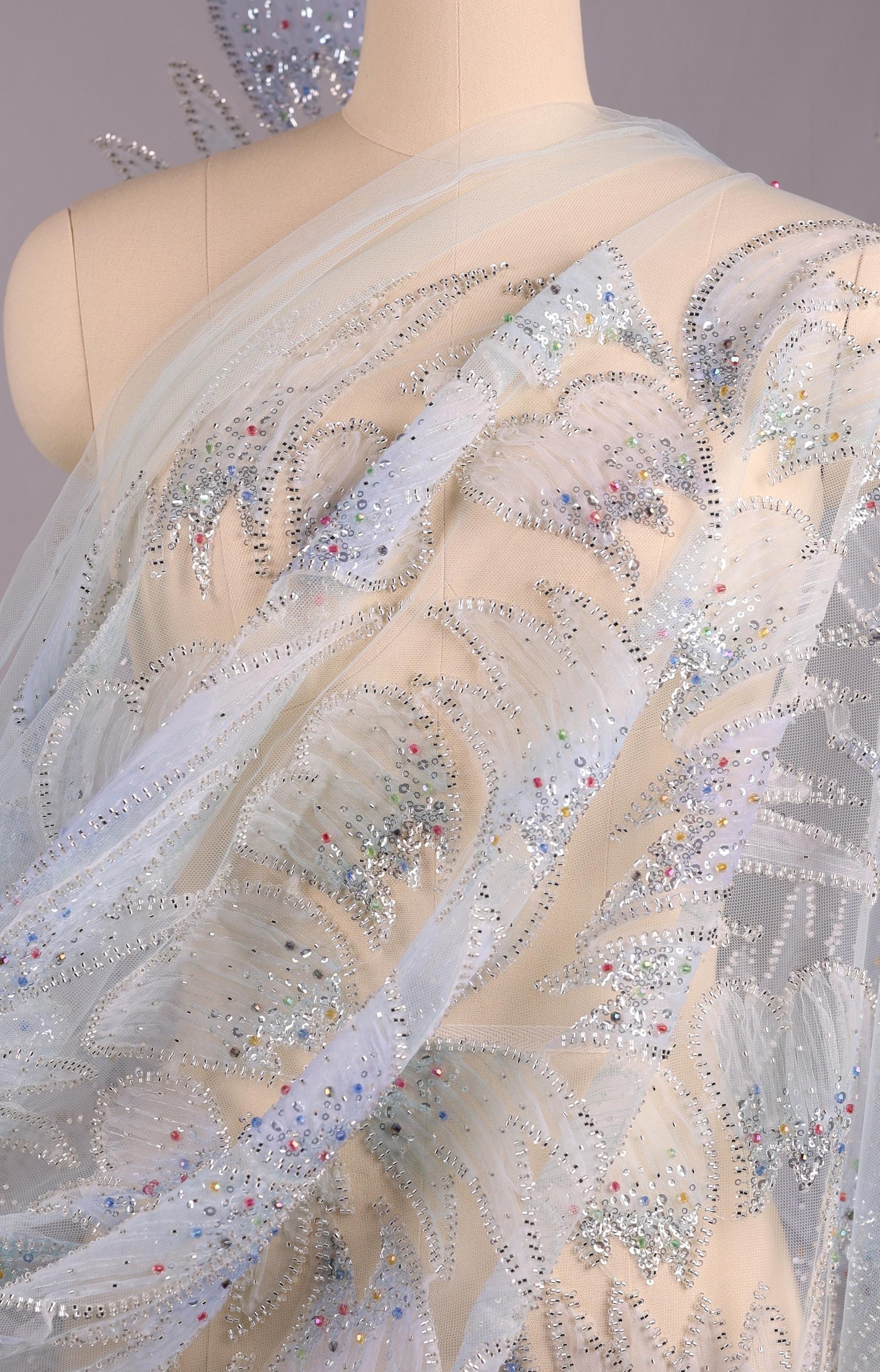 a mannequin wearing a white dress with a sheer, sheer-like fabric and a floral pattern. The dress appears to be made of sheer white fabric with silver embellishments, such as sequins or rhinestones, adding a touch of sparkle and elegance to the design.