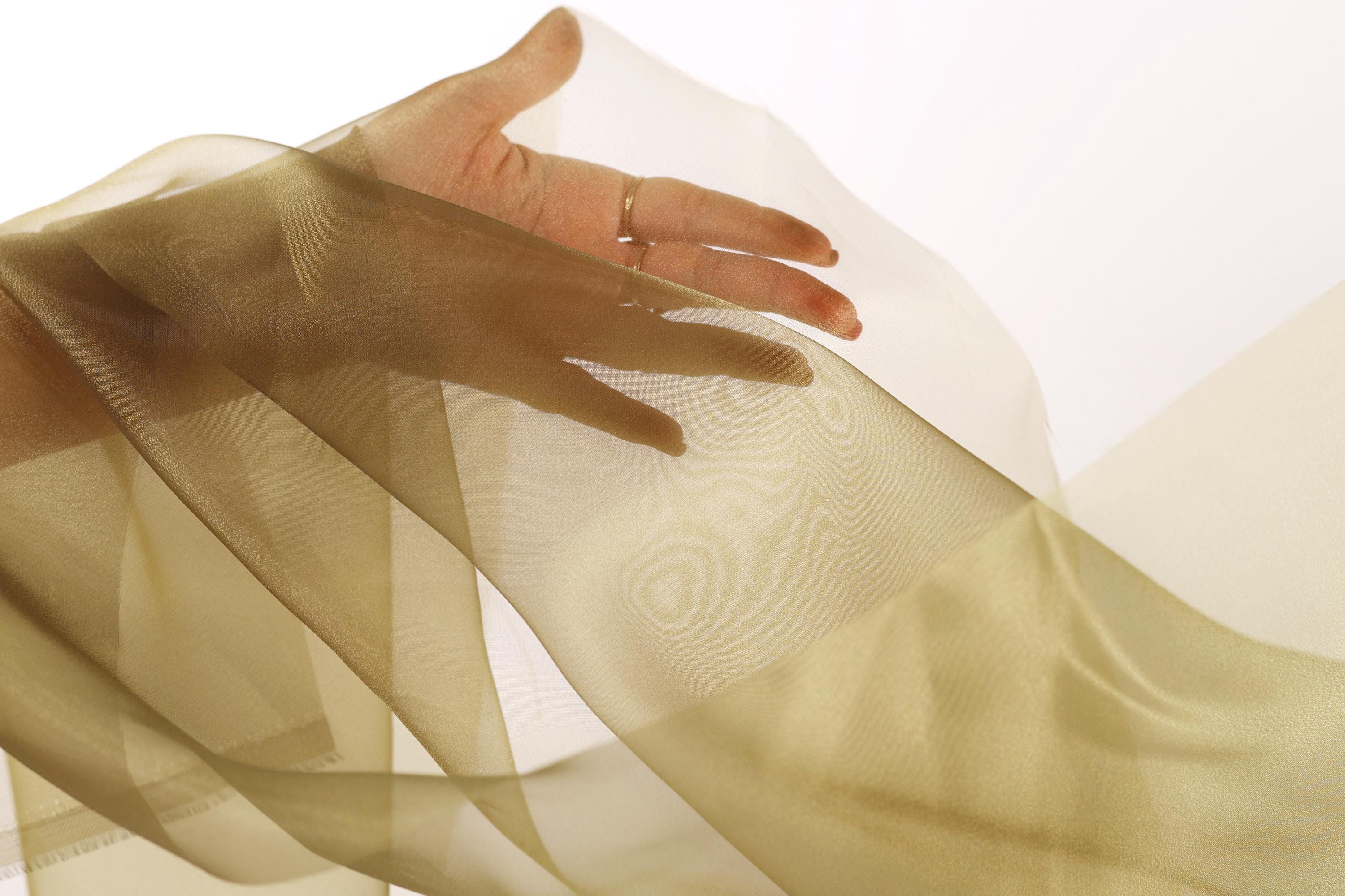 A hand is holding a piece of light brown fabric, which appears to be a sheer or sheer-like material, against a white background.