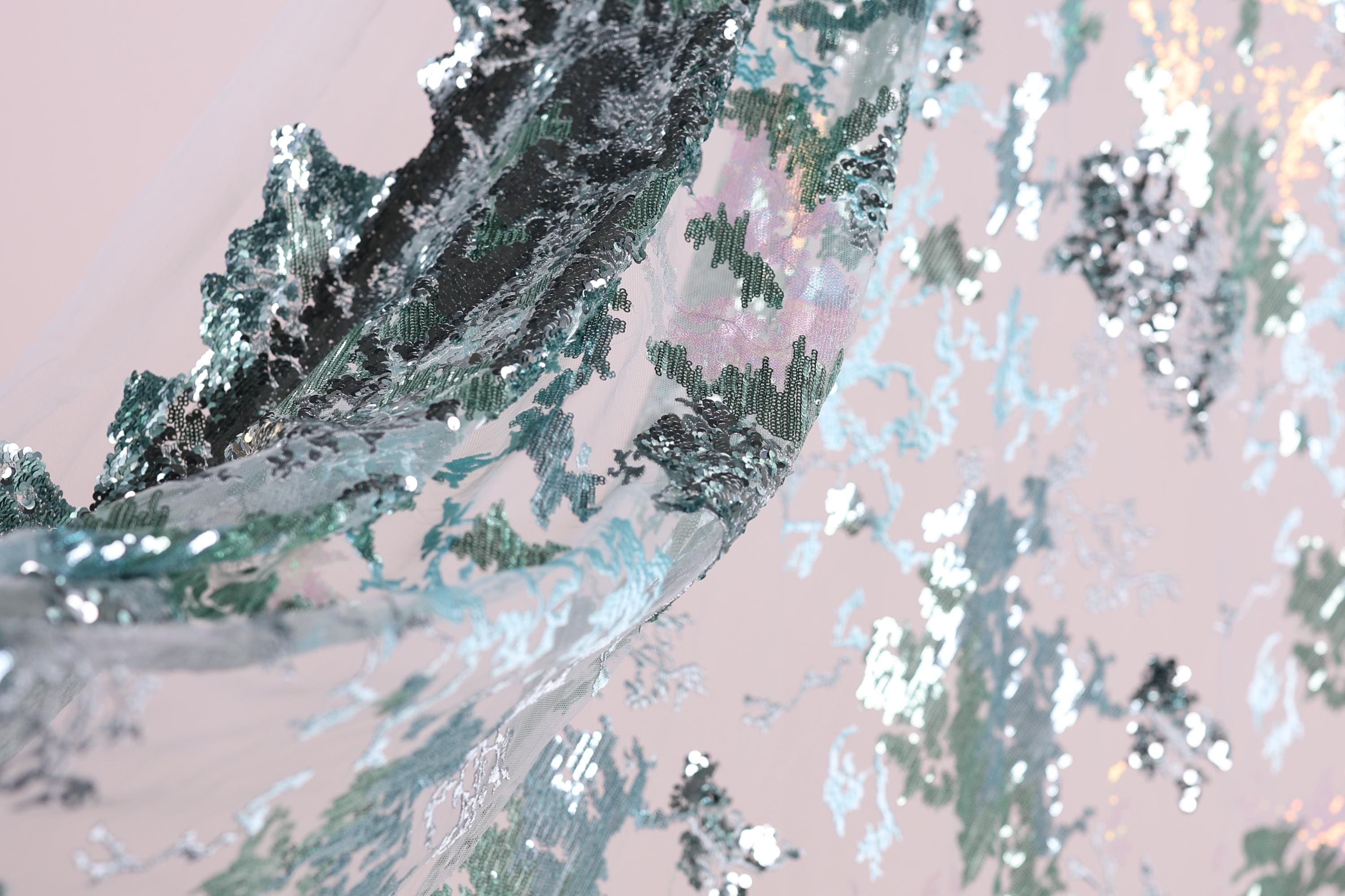 The image is a close-up of a fabric with a metallic, sequined texture. The fabric appears to be a sheer material, with a pattern of green and silver sequins that create a visually striking and intricate design. The fabric is not a single piece but rather a part of a larger fabric, as suggested by the background that is a light pink color. The image does not contain any discernible text or countable objects, and the focus is on the detailed and textured appearance of the fabric.