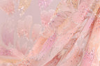a close-up view of a delicate, pink and white fabric with a floral pattern. The fabric appears to be made of sheer material, giving it a soft and delicate appearance. The background is blurred, drawing focus to the fabric and its intricate details.