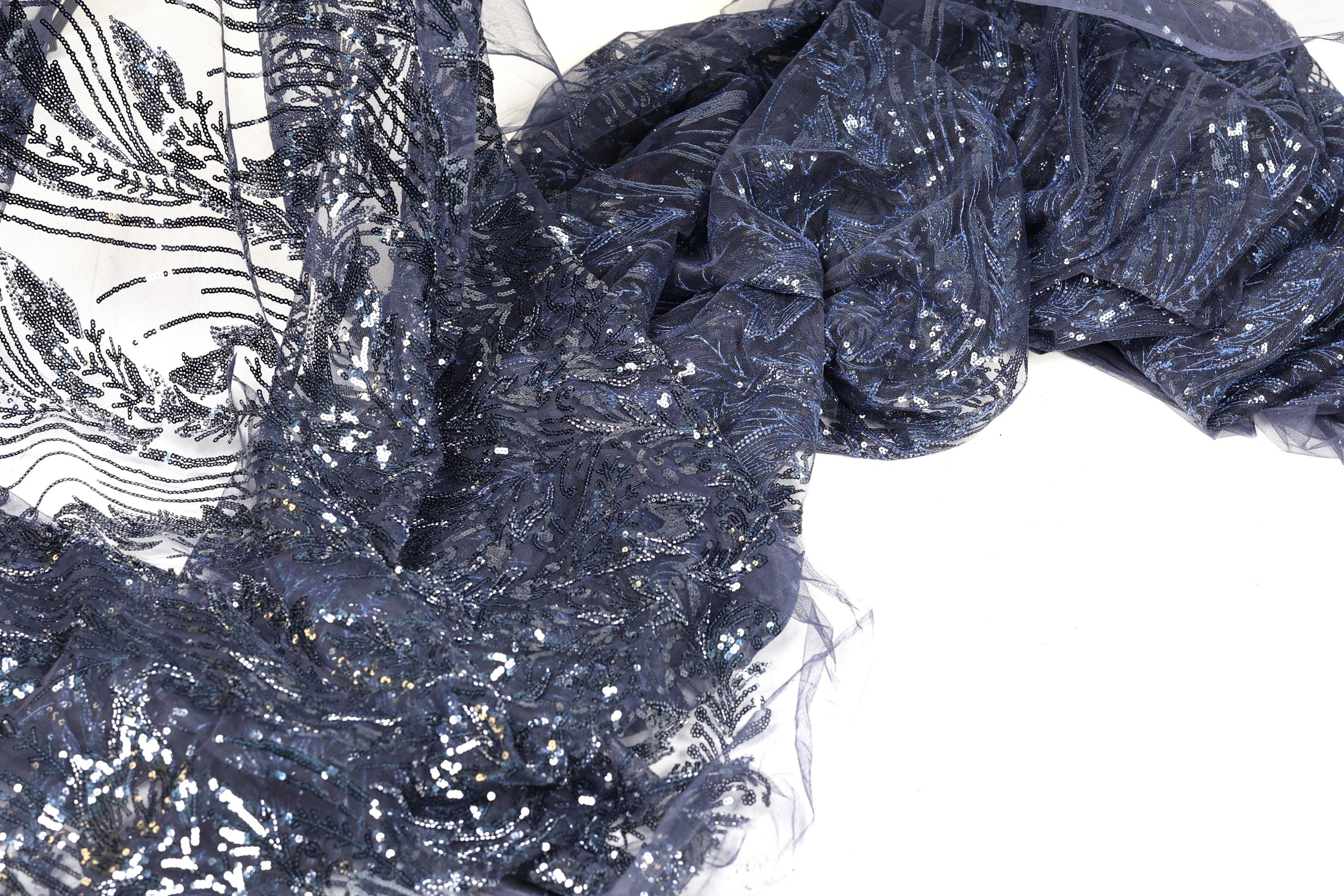 a close-up view of a fabric with a dark blue, glittery, and sequined pattern. The fabric appears to be a type of lace or sheer material, with a textured and shiny appearance.