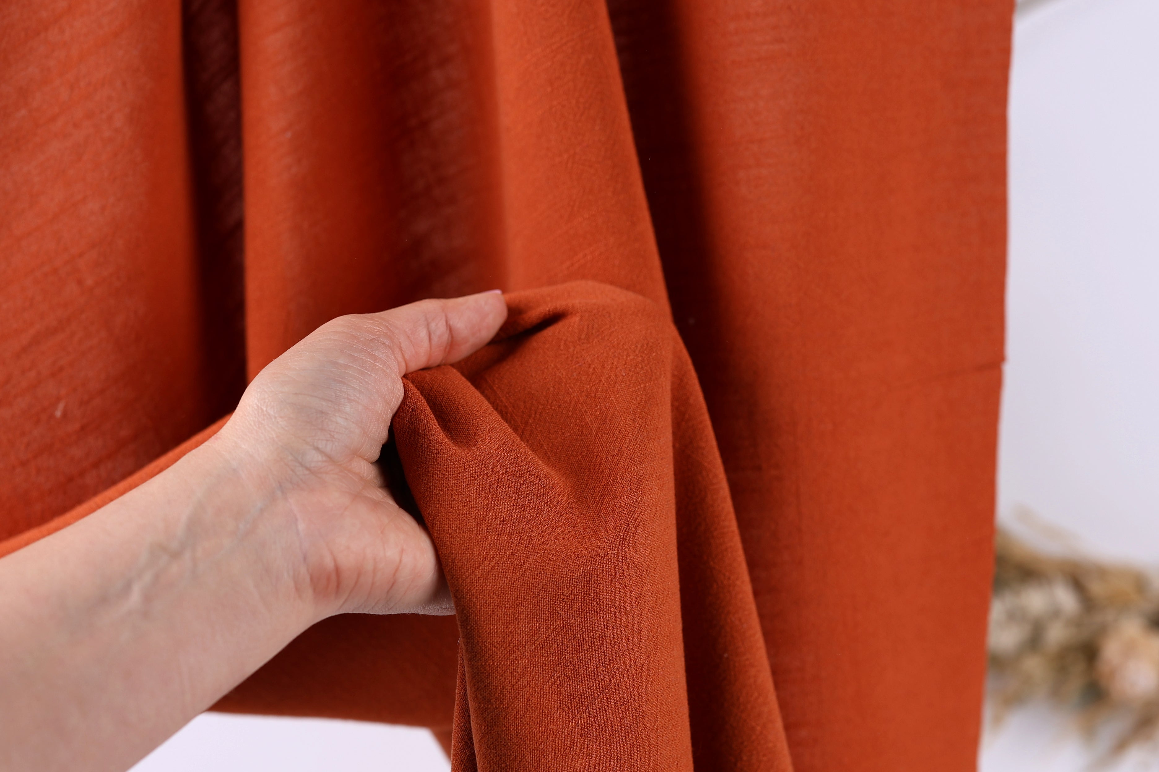 A hand is holding a piece of orange fabric, possibly a curtain or garment, in front of a red curtain or fabric background.