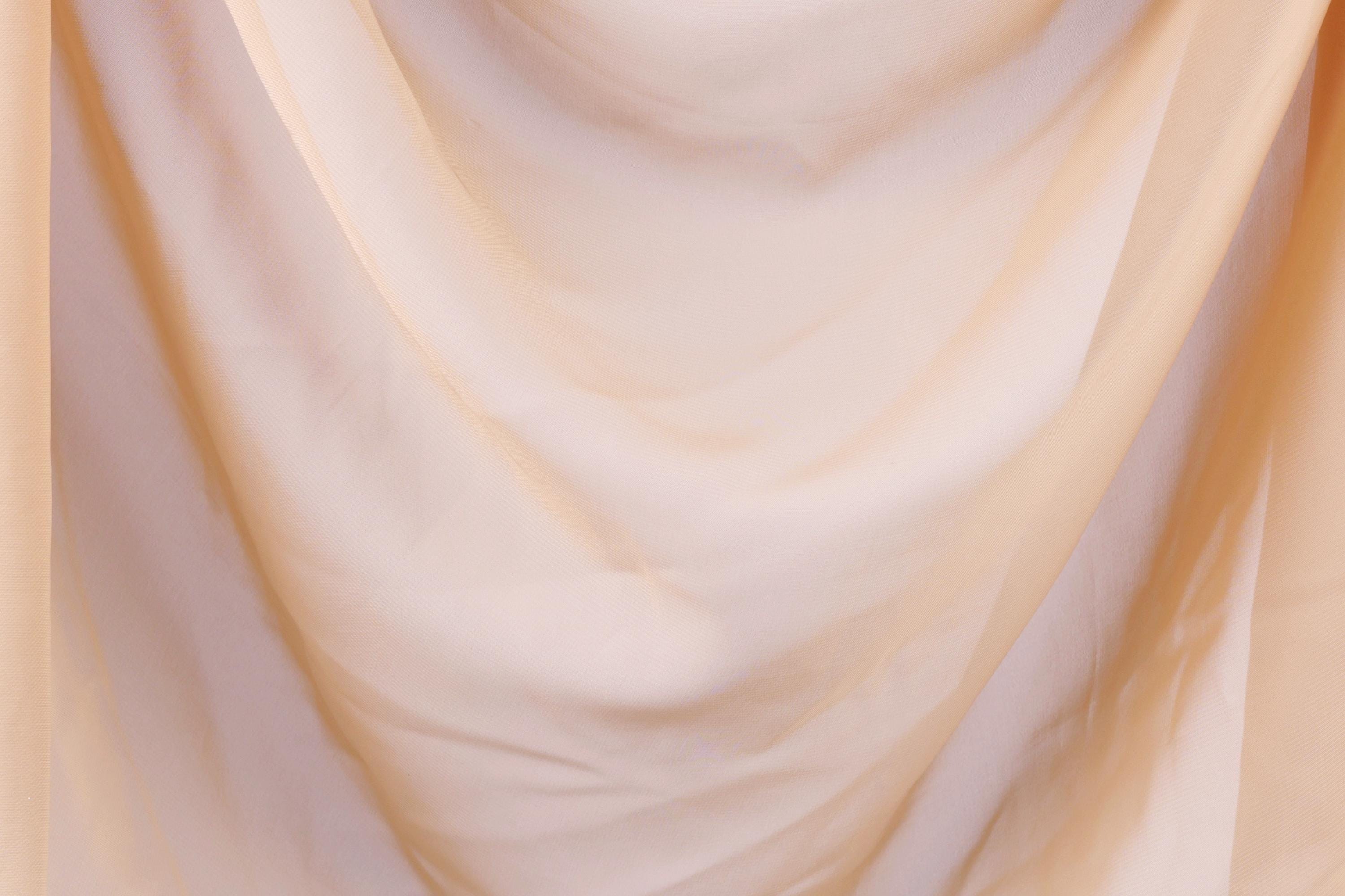 The image is a close-up of a soft, flowing fabric, possibly a sheer or sheer-like material, with a light peach or pale yellow color. The fabric appears to be made of a delicate, sheer material, which gives it a delicate and airy appearance.