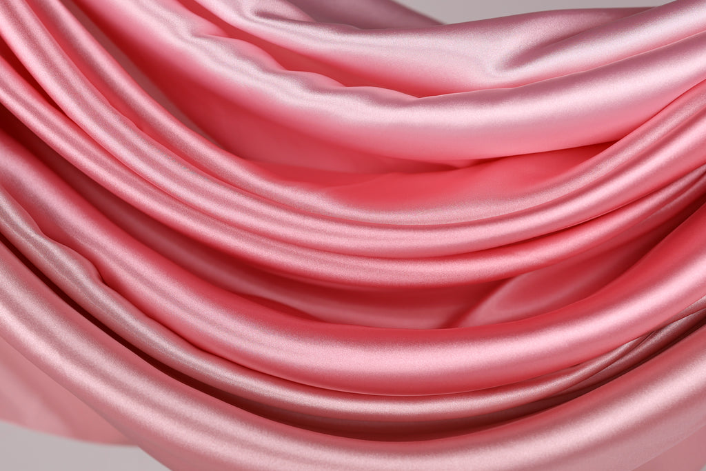 The image is a close-up of a soft, flowing pink satin fabric, with its folds and creases creating a sense of depth and texture.