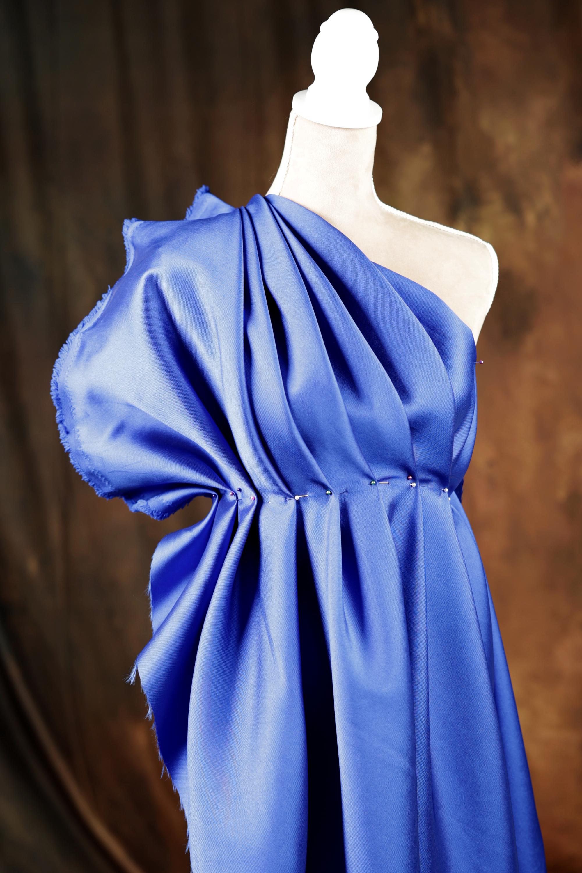 A blue one-shoulder dress with a draped, flowing silhouette is displayed on a mannequin.