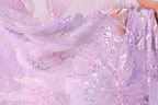 A close-up view of a delicate, shimmering fabric with a purple and pink color scheme, adorned with small, multicolored beads or sequins that create a sparkling effect.