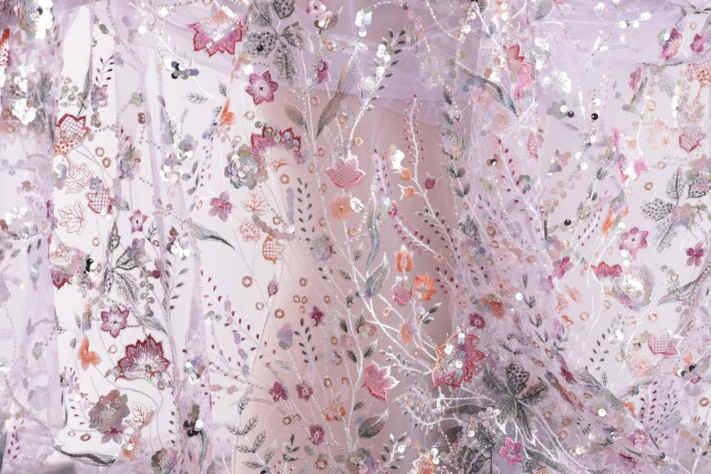 The image is a close-up view of a delicate, intricately embroidered fabric with a floral pattern. The fabric appears to be made of sheer material, giving it a soft and delicate appearance. The embroidered flowers and leaves are arranged in a way that creates a visually appealing and intricate design.