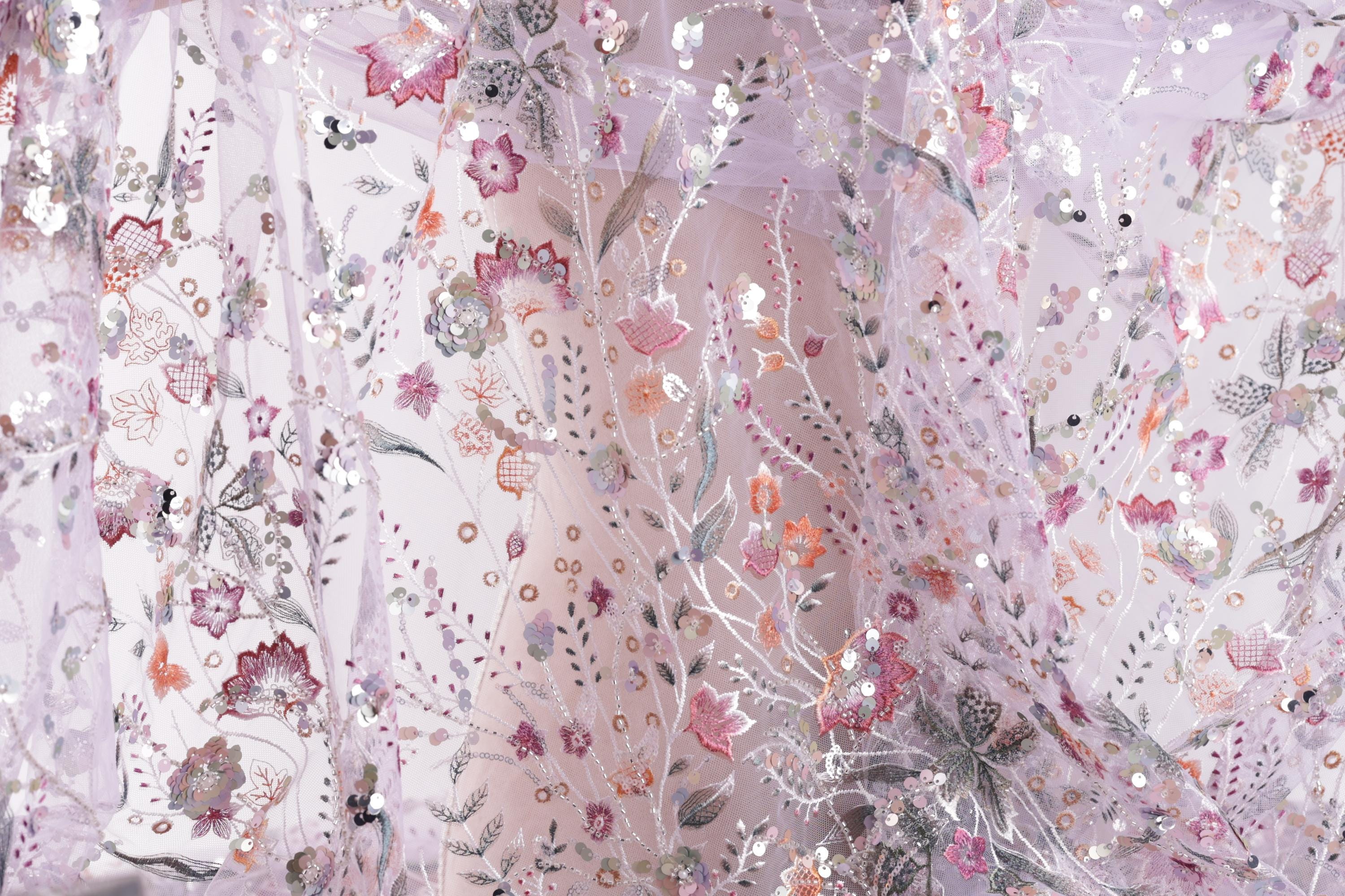 The image is a close-up view of a delicate, intricately embroidered fabric with a floral pattern. The fabric appears to be made of sheer material, giving it a soft and delicate appearance. The embroidered flowers and leaves are arranged in a way that creates a visually appealing and intricate design.