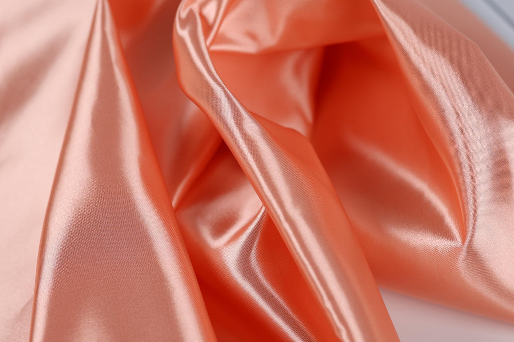 a close-up view of a soft, shiny fabric with a peach-colored hue. The fabric appears to be made of satin or a similar smooth material, and it has a slightly wrinkled texture, giving it a flowing and elegant appearance.
