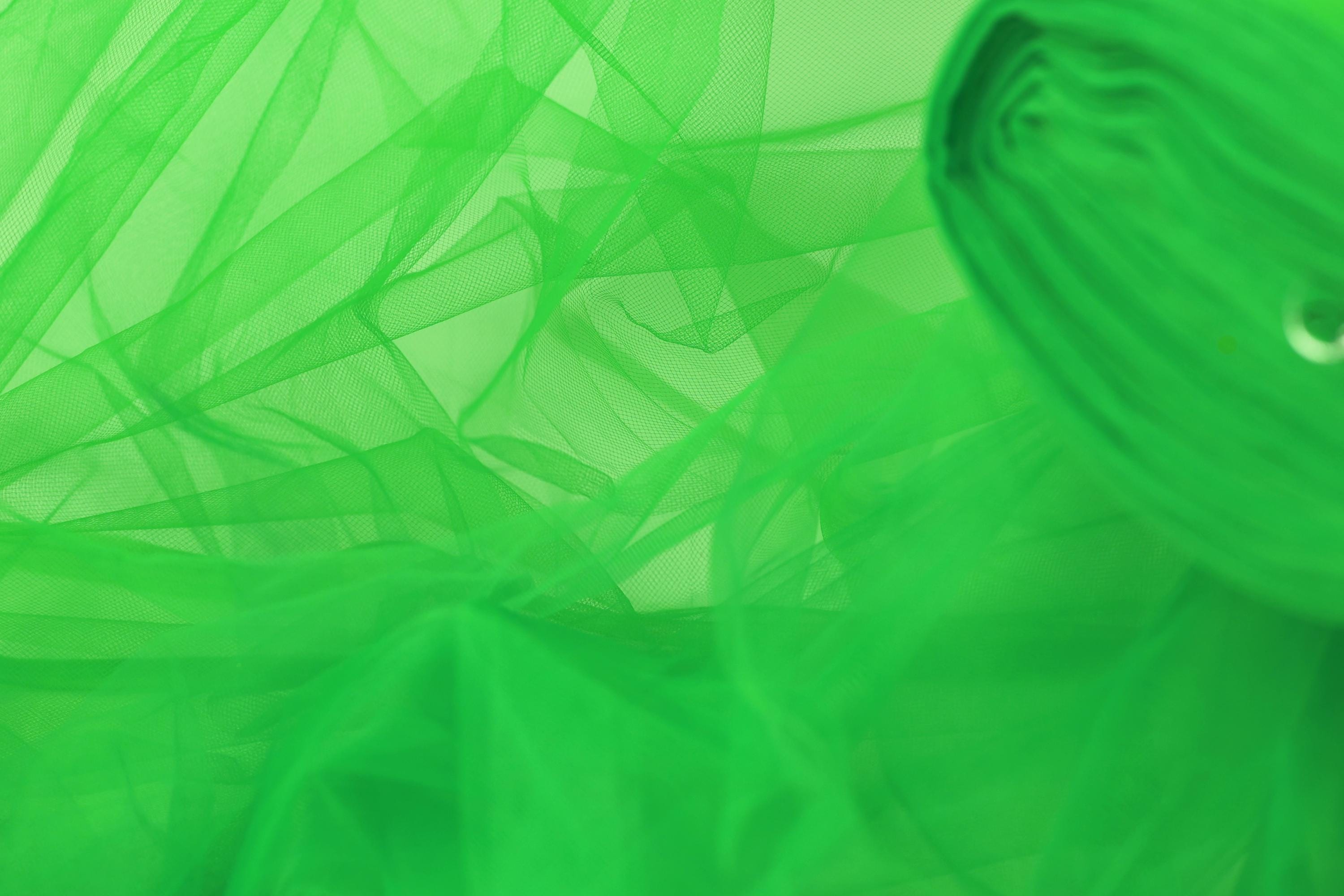 The image is a close-up of a green, translucent fabric, possibly a type of tulle or chiffon, with a blurred, abstract background.