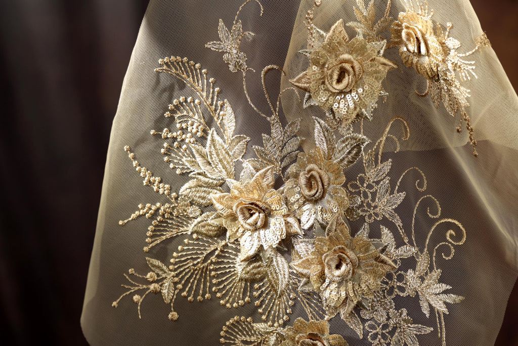 a close-up view of a delicate, intricately embroidered fabric with a floral design. The fabric appears to be a sheer material, with gold-colored flowers and leaves adorning the surface. The embroidery is detailed and appears to be handcrafted, showcasing the skill and artistry involved in creating this piece of textile art.