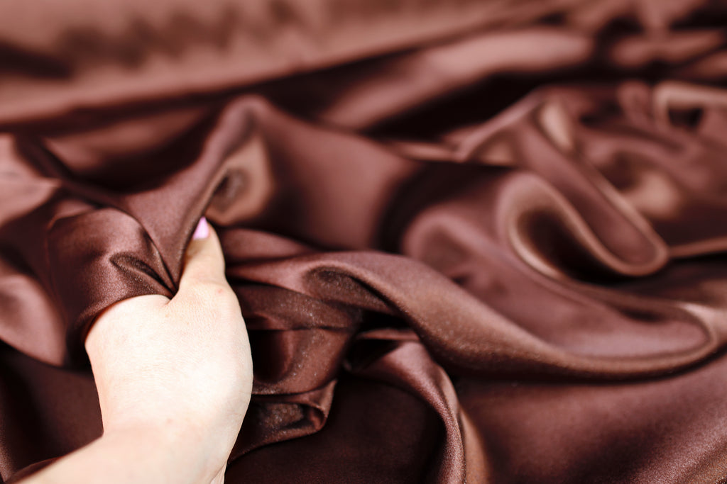A hand is holding a piece of brown satin fabric, which appears to be a close-up view of the material.