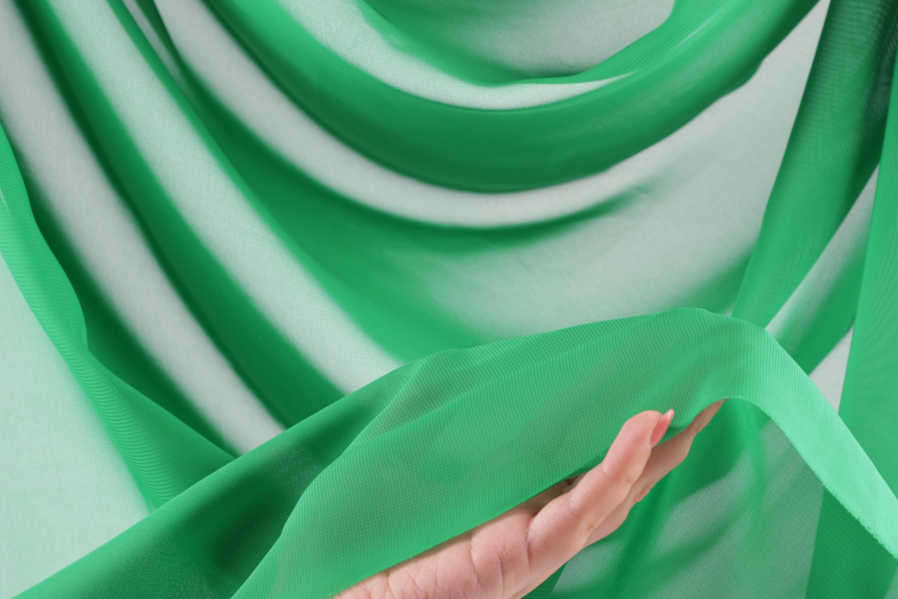 A hand is holding a piece of green fabric, which appears to be a sheer or sheer-like material. The fabric is draped over the hand, creating a soft and flowing appearance.