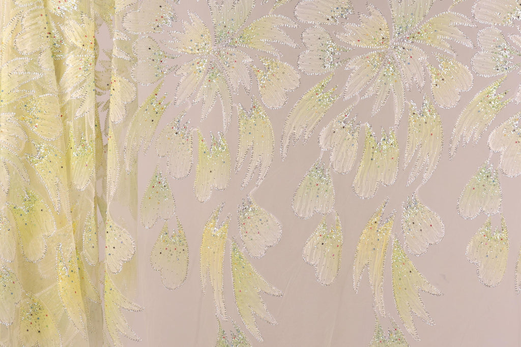 a close-up view of a fabric with a floral pattern, including yellow and white flowers and leaves. The fabric appears to be made of a shiny, glittery material, giving it a sparkly and delicate appearance.