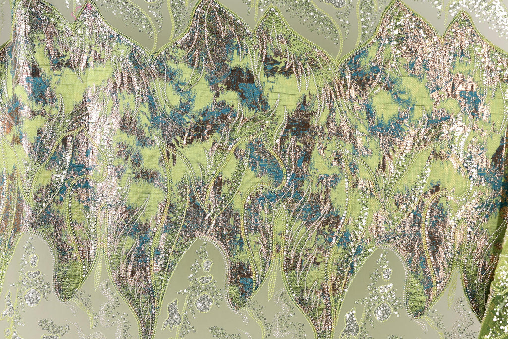 The image is a close-up of a fabric with a vibrant, multicolored pattern. The fabric appears to be made of a shiny, metallic material, with a mix of green, blue, and silver colors. The pattern is intricate and detailed, with various shapes and textures that create a visually striking and dynamic design.