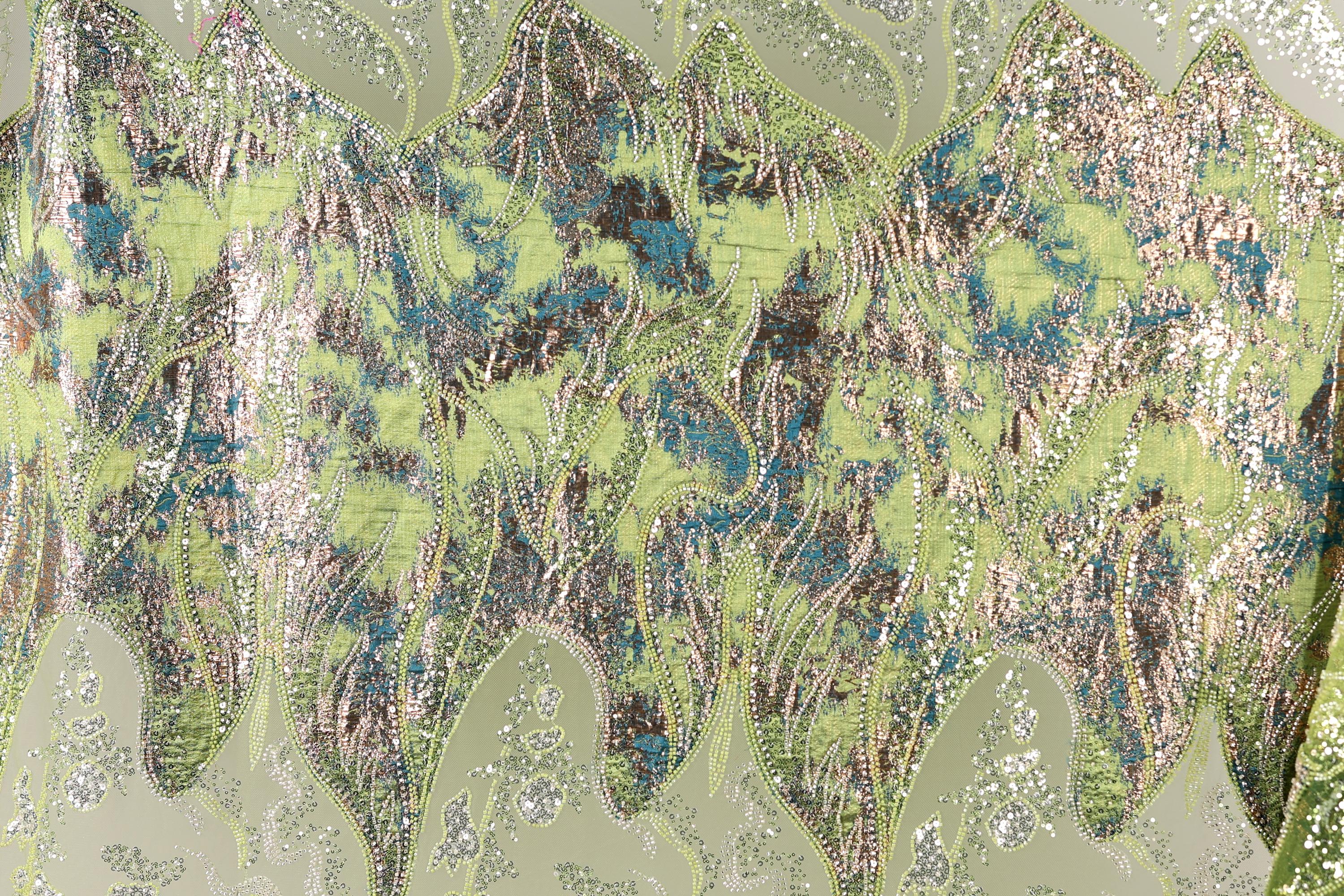 The image is a close-up of a fabric with a vibrant, multicolored pattern. The fabric appears to be made of a shiny, metallic material, with a mix of green, blue, and silver colors. The pattern is intricate and detailed, with various shapes and textures that create a visually striking and dynamic design.