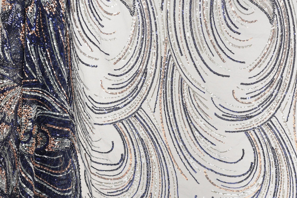 a close-up view of a fabric with a repeating pattern of sequin designs in shades of blue, white, and gold. The pattern consists of curved lines and swirls, creating a visually striking and intricate design.