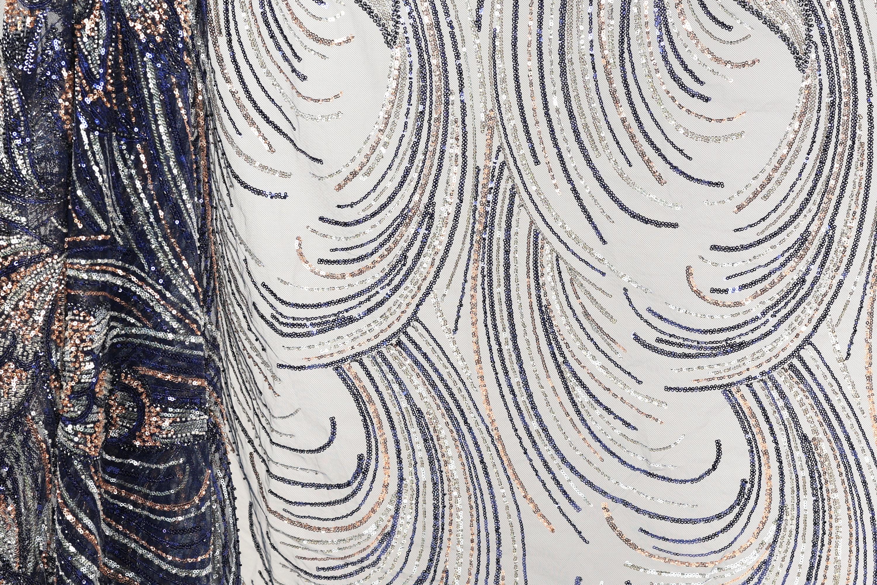 a close-up view of a fabric with a repeating pattern of sequin designs in shades of blue, white, and gold. The pattern consists of curved lines and swirls, creating a visually striking and intricate design.