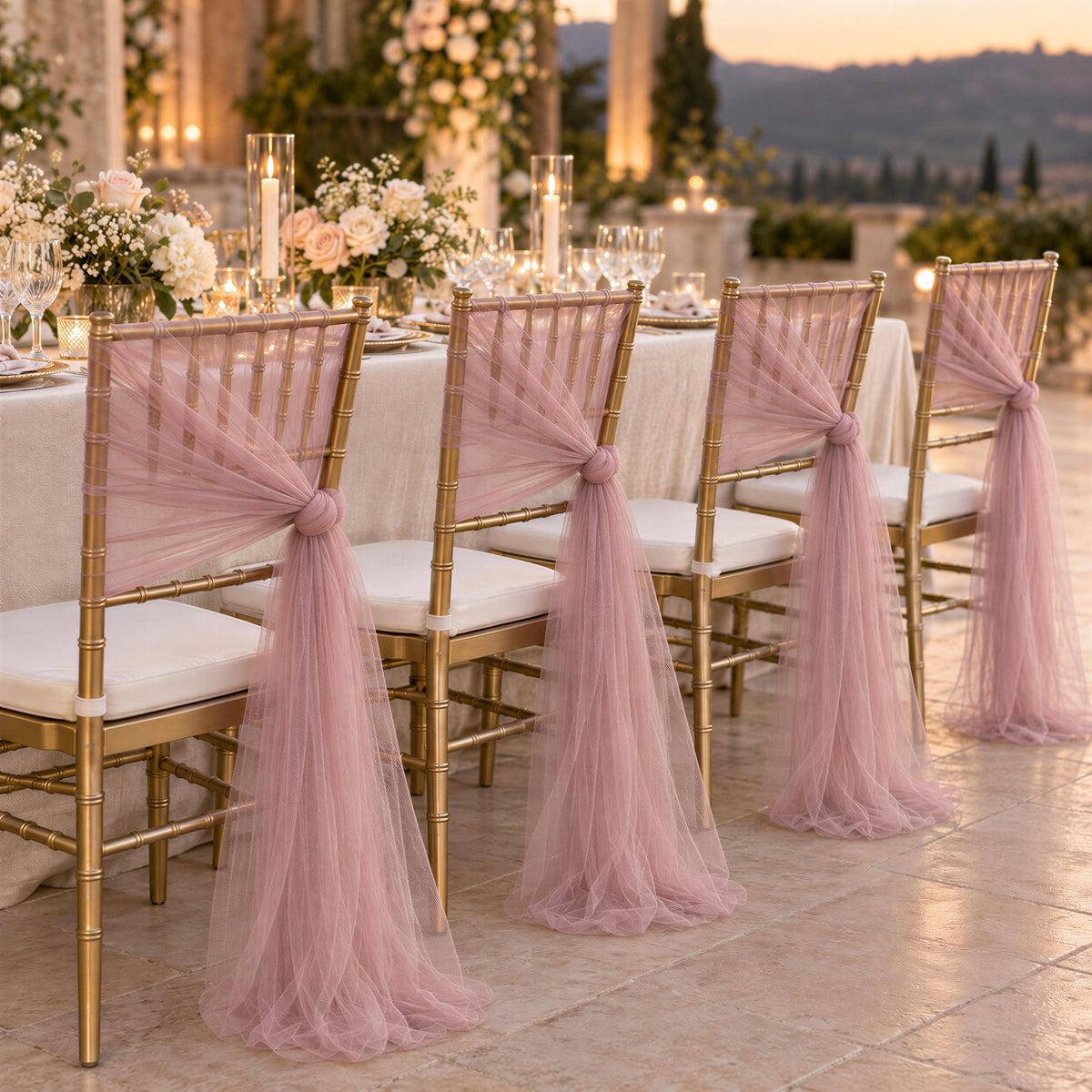a beautifully decorated outdoor wedding reception with a table set for four guests. The chairs are adorned with pink tulle fabric draped over the backrests, creating an elegant and romantic atmosphere. The table is set with white tablecloths, candles, and floral centerpieces, suggesting a formal and sophisticated event.