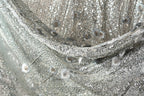 The image is a close-up of a fabric with a silver and white pattern, featuring small silver and white sequins or beads.