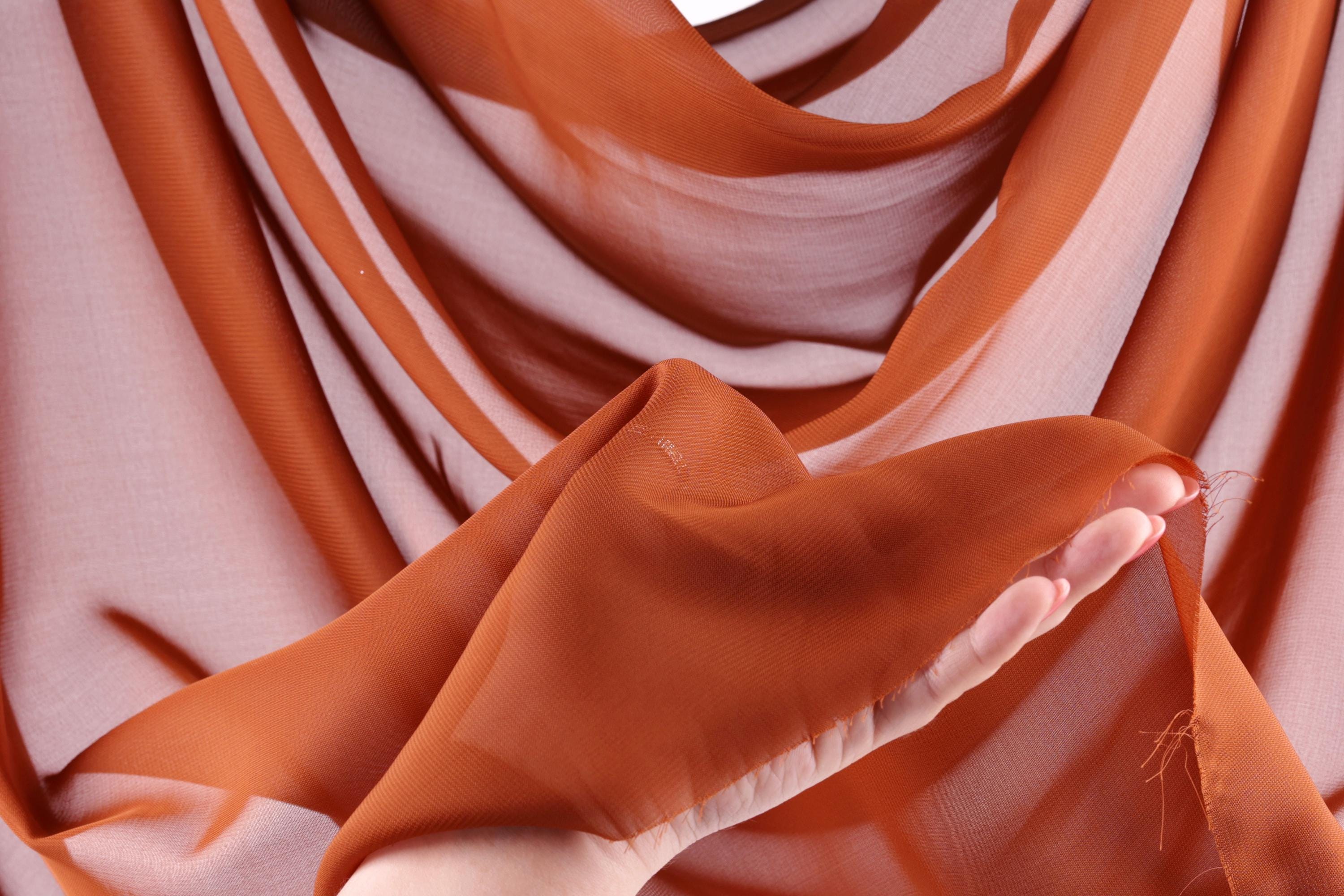A hand is holding a piece of orange fabric, which appears to be a scarf or garment. The fabric has a textured and wrinkled appearance, suggesting it is made of a soft, natural material.