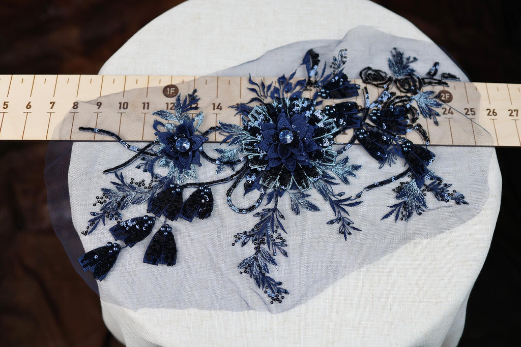 a close-up view of a piece of fabric with a floral design, including blue and black sequins and tassels. The fabric is placed on a white surface, and a ruler is visible in the background.