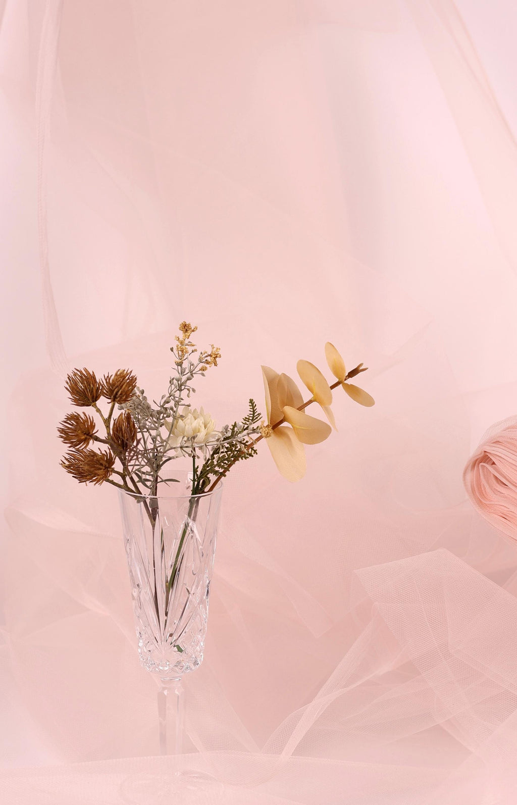 A clear glass vase filled with a variety of flowers, including dried flowers and a single yellow flower, sits on a pink background.