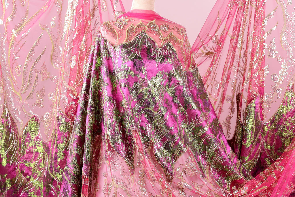 a vibrant, intricately designed fabric with a mix of pink, green, and gold colors. The fabric appears to be a sheer or sheer-like material, with a pattern of leaves and flowers that adds a touch of nature to the design. The fabric is displayed in a way that showcases its details and patterns, making it an eye-catching and visually appealing piece.