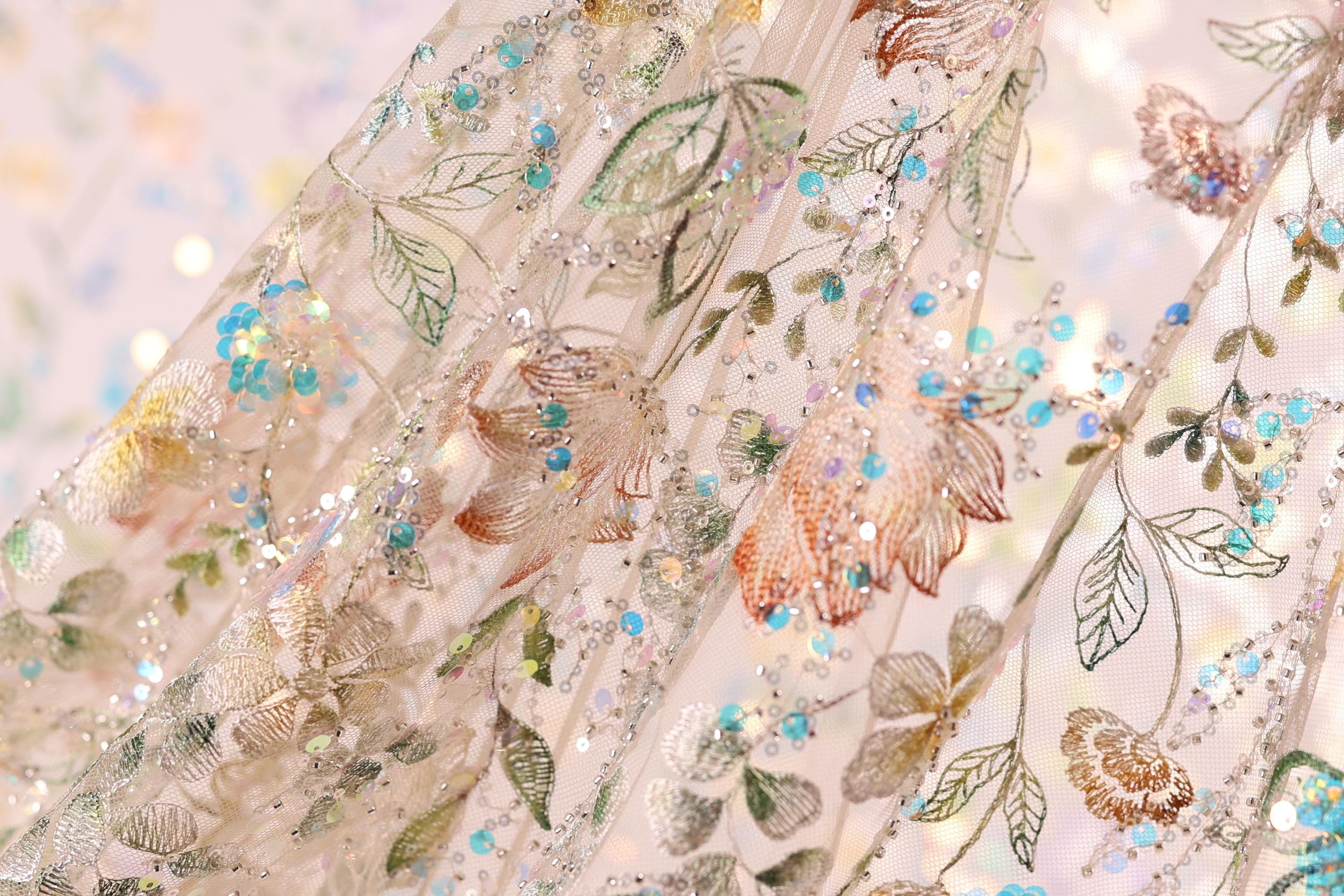 The image is a close-up of a delicate, intricately embroidered fabric with a floral pattern. The fabric is adorned with small, colorful beads and sequins that add a touch of sparkle and elegance to the design. The close-up view allows for a detailed examination of the fabric's texture and the intricate patterns created by the embroidery.