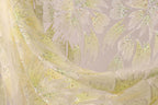 a close-up view of a delicate, sheer fabric with a floral pattern. The fabric appears to be made of a light-colored material, possibly silk or chiffon, with a shimmering, sparkly texture. The pattern on the fabric consists of various shades of green and yellow, creating a visually appealing and intricate design. The fabric is spread out, allowing for a clear view of its details and textures.