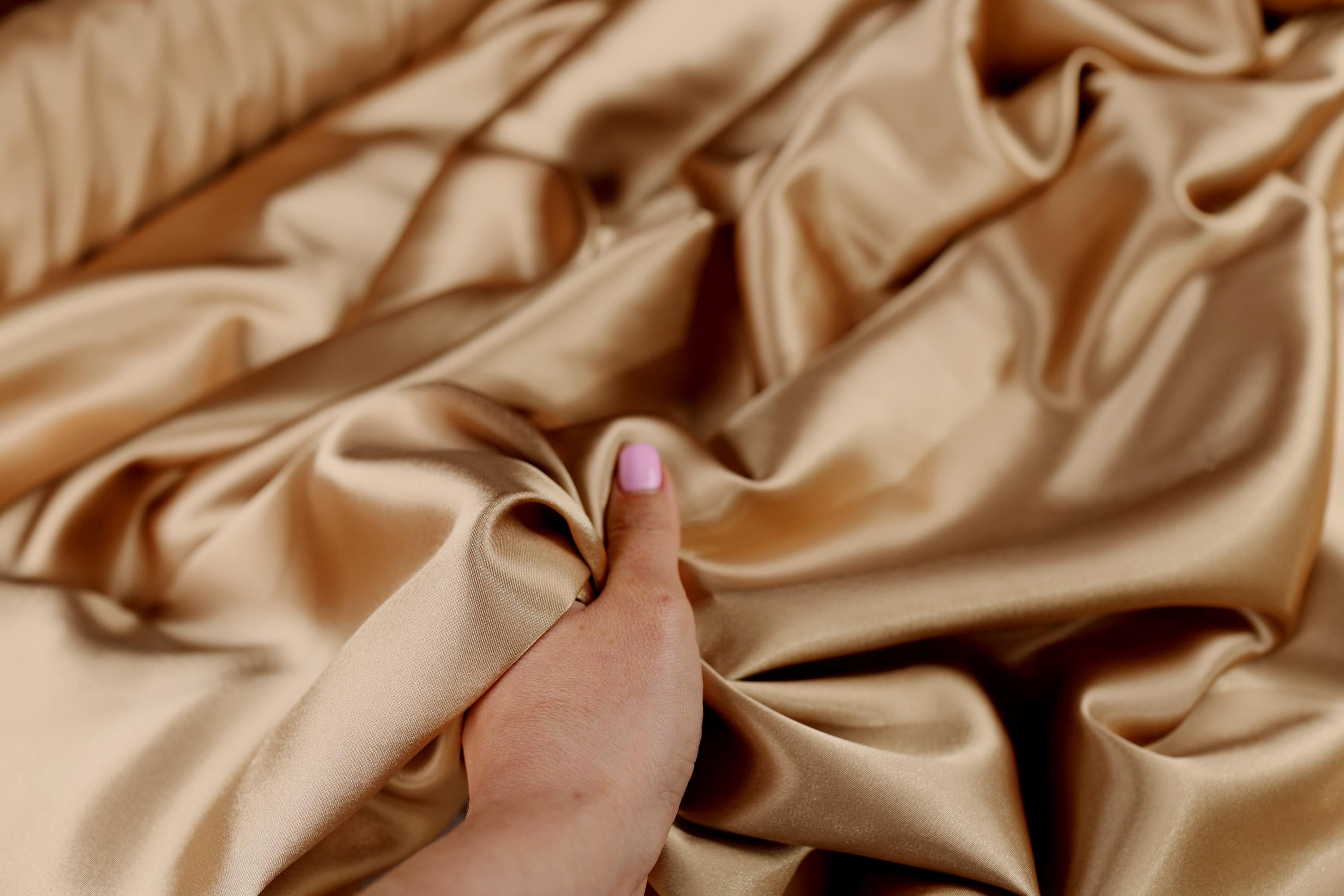 A hand is holding a piece of gold satin fabric, which appears to be soft and smooth.
