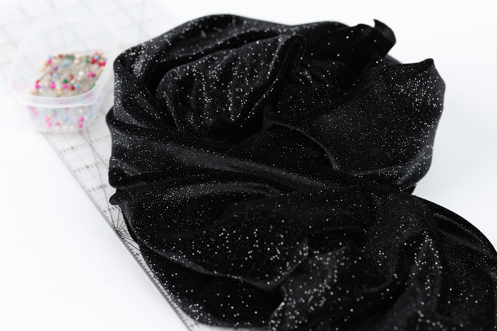 a black fabric with a glittery texture, placed on a white surface next to a clear container filled with small, multicolored pins.