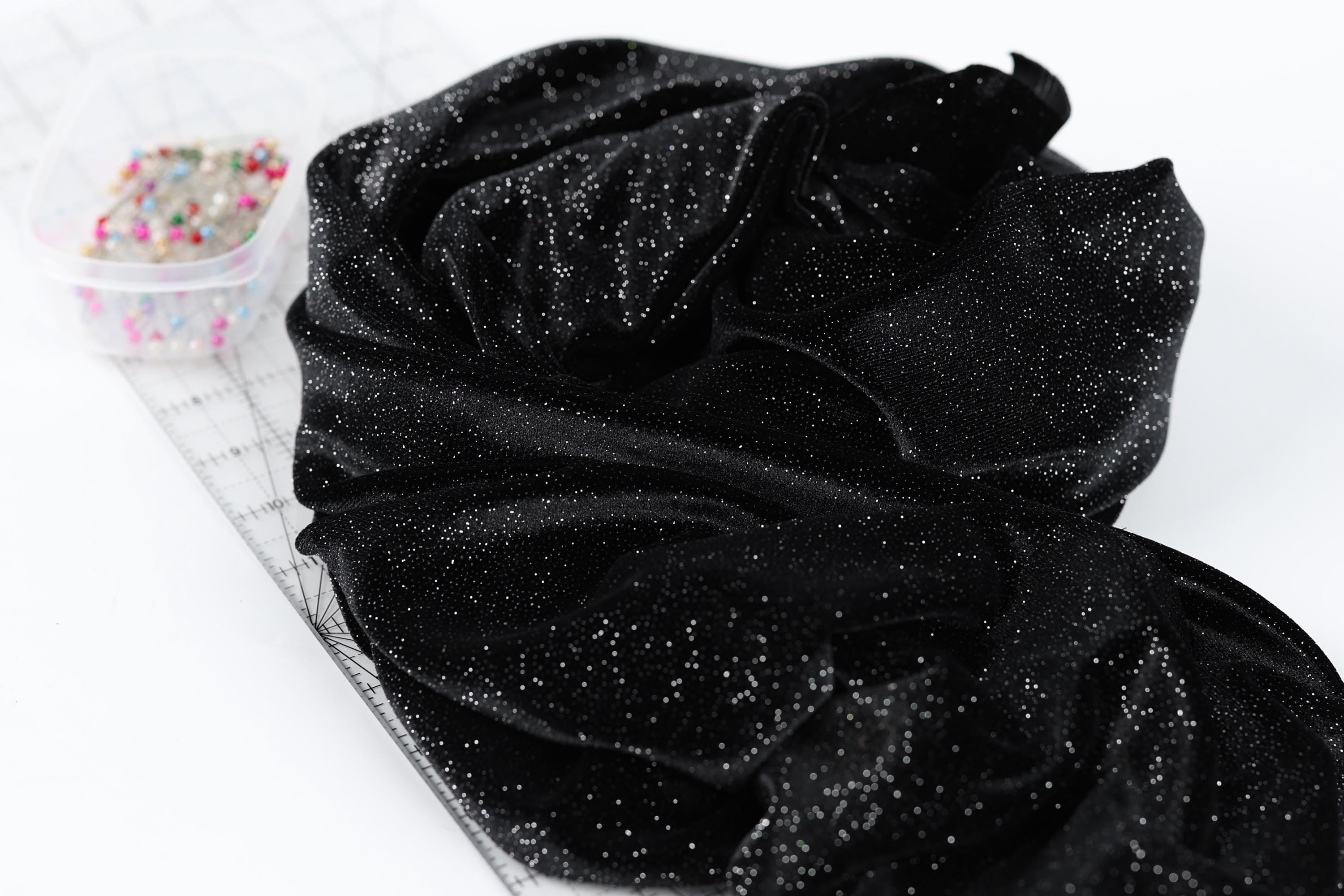 a black fabric with a glittery texture, placed on a white surface next to a clear container filled with small, multicolored pins.