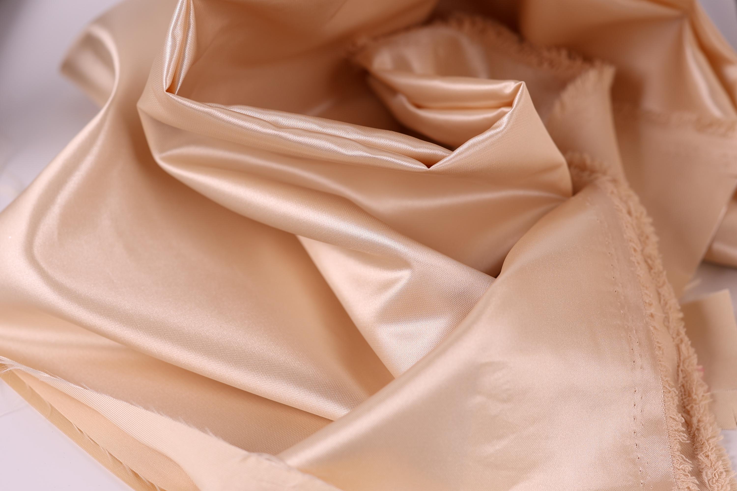 A close-up view of a soft, light-colored fabric, possibly silk or satin, with a smooth and shiny texture.