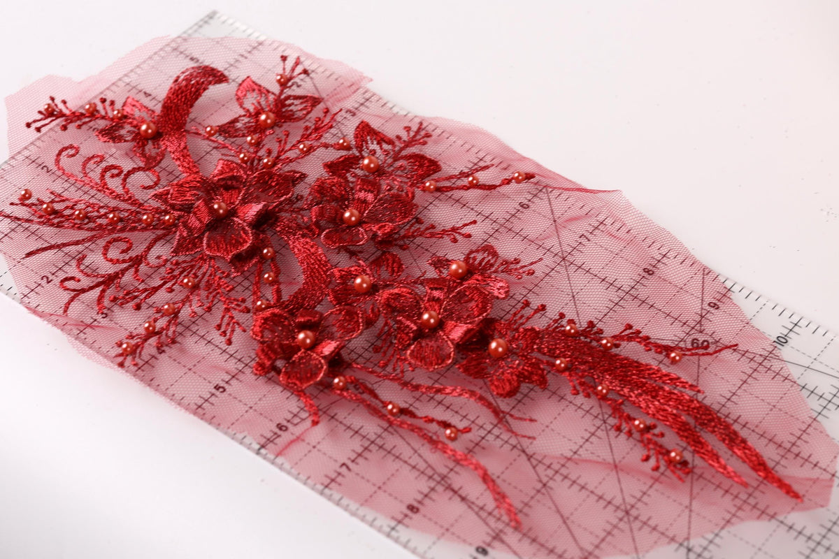 a close-up view of a red floral lace piece with intricate details, including red flowers and beads, placed on a pink background.