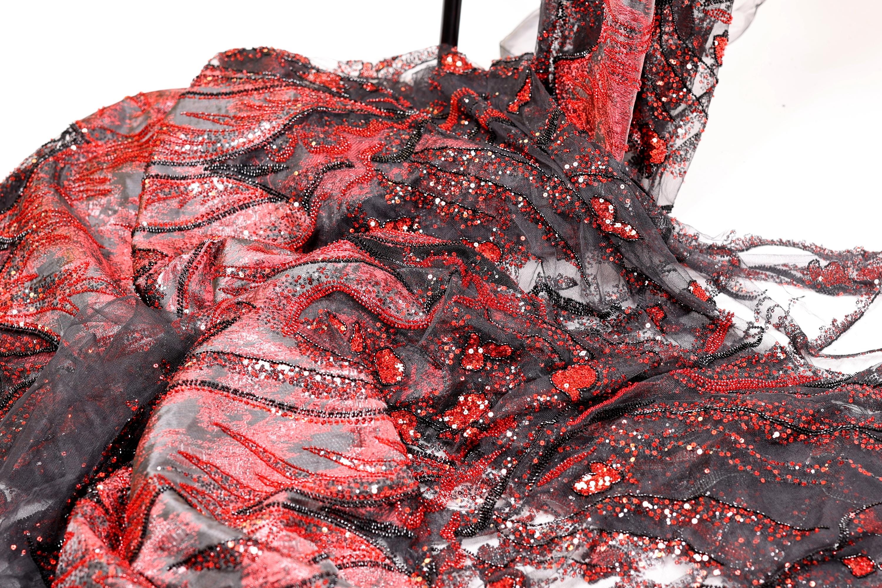 a close-up view of a fabric with a striking pattern of red and black sequins and beads. The fabric appears to be a sheer material, with a mix of black and red colors. The pattern is intricate and detailed, with the sequins and beads adding a touch of sparkle and glamour to the fabric.
