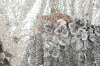 a close-up view of a fabric with a floral pattern, including various shades of gray and white flowers and leaves. The fabric appears to be made of sequins or beads, adding a touch of sparkle and elegance to the design.