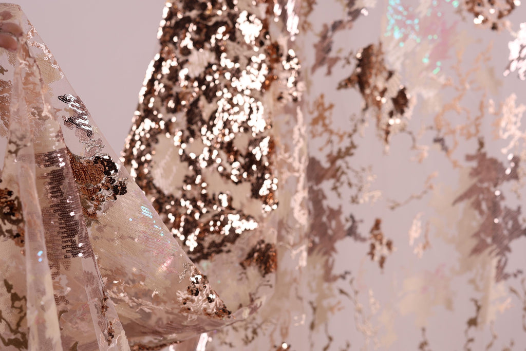 a close-up view of a fabric with a metallic, sequined texture. The fabric appears to be a sheer material, with a pattern of small sequins and beads that create a shimmering effect. The background of the image is blurred, drawing focus to the fabric and its intricate details.