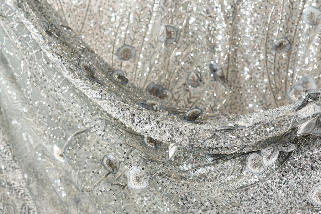 The image is a close-up of a fabric with a silver sequin pattern, featuring a mix of small and larger sequins, as well as some floral or leaf-like elements. The fabric appears to be made of a shiny, metallic material, giving it a sparkling and luxurious appearance.